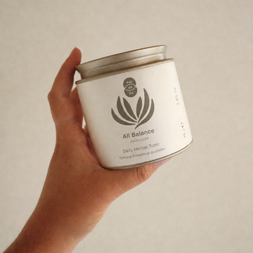 A hand holding a white cylindrical tin of All Balance Herbal Tonic against a neutral light gray background.