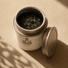 An open round metal tea tin filled with loose-leaf tea, with its lid resting beside it on a warm beige surface. The tin has a white label with a plant /flower logo and the brand name All Of Us and the Product name All Balance. 