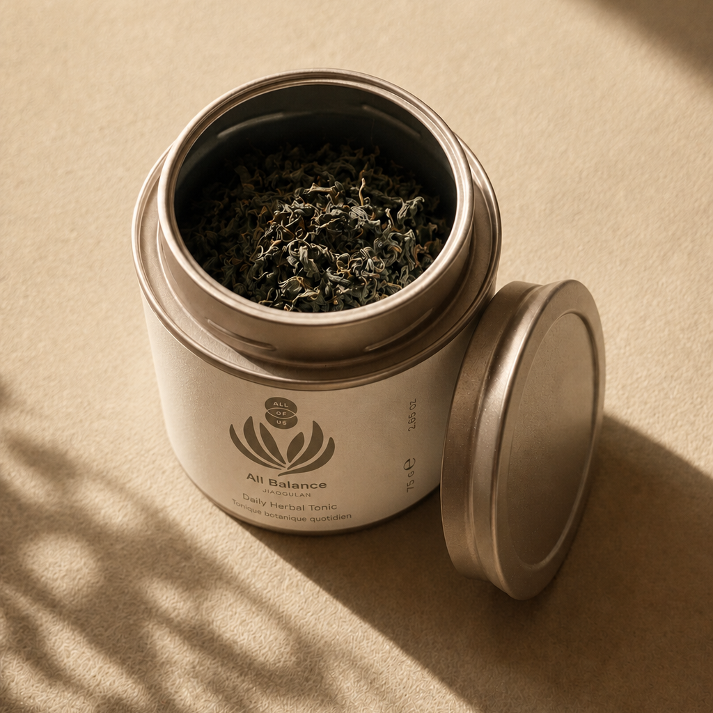 An open round metal tea tin filled with loose-leaf tea, with its lid resting beside it on a warm beige surface. The tin has a white label with a plant /flower logo and the brand name All Of Us and the Product name All Balance. 