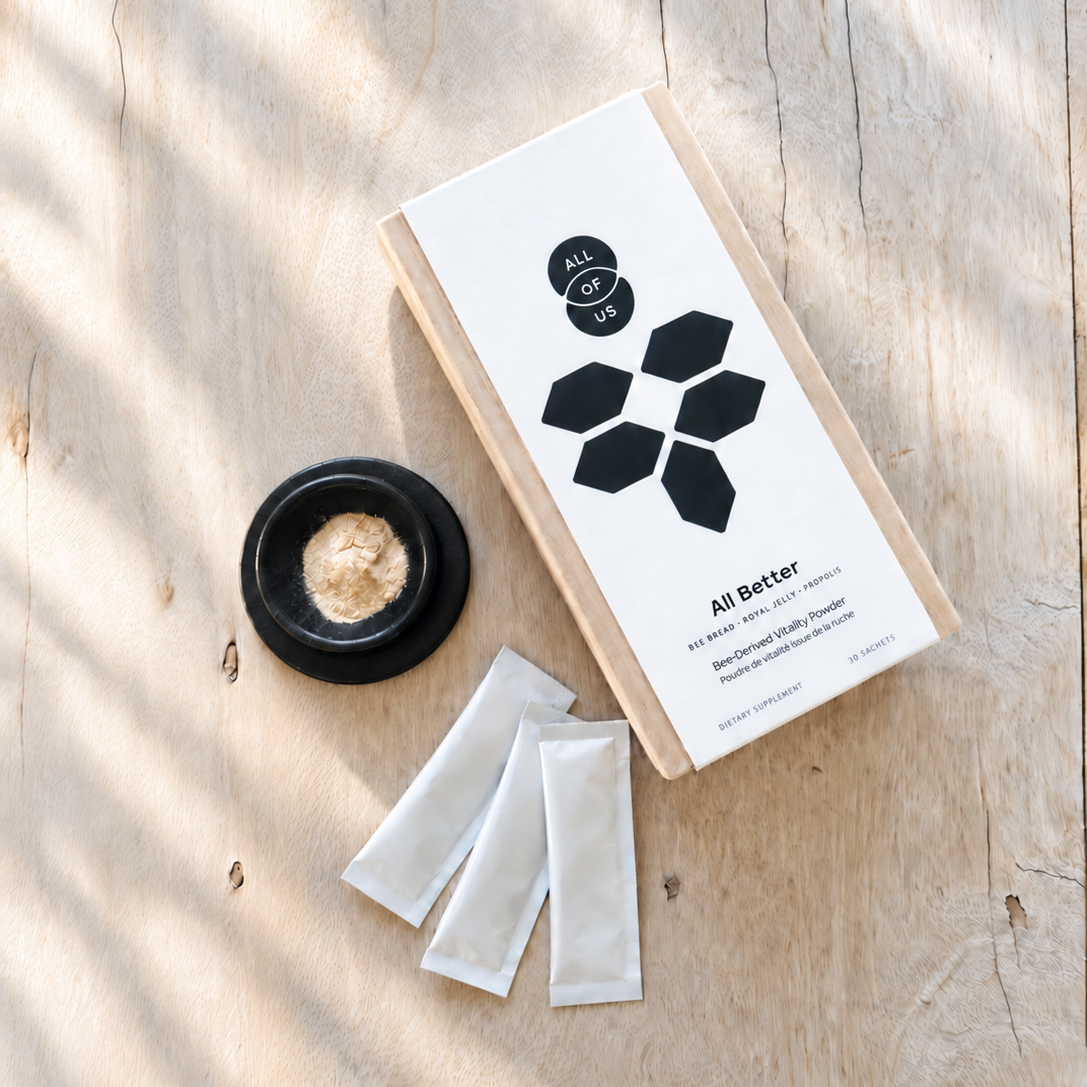 A flat-lay product shot of 'All Better' herbal tea by All In One on a light wood background with soft leaf shadows. The set includes a natural wood box with a white label, three white sachets, and a small black dish containing the powder.