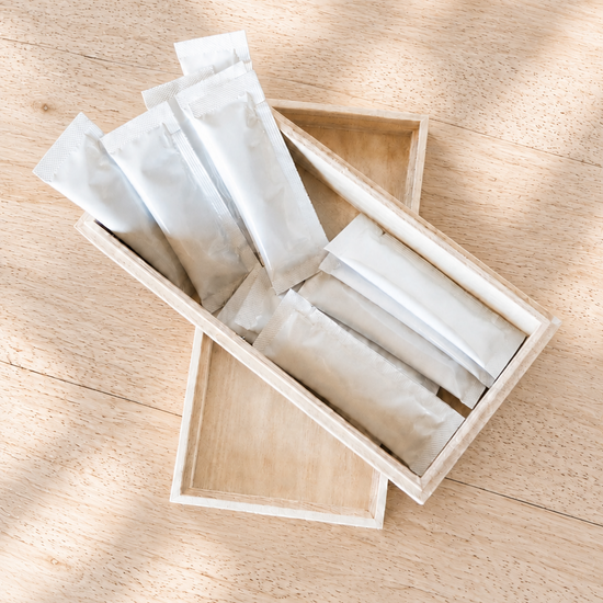 Several small white sachets or packets arranged inside an open light wood tray on a pale wood surface with soft dappled shadows.