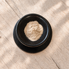 A small mound of light beige powder sits in a black ceramic bowl with a matching saucer, placed on a light wood surface with soft leaf shadows.