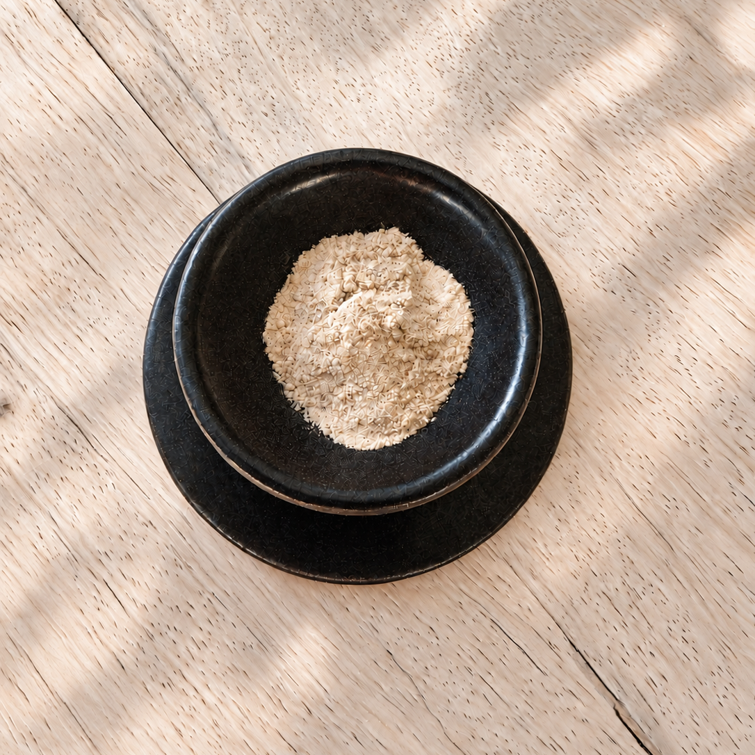 A small mound of light beige powder sits in a black ceramic bowl with a matching saucer, placed on a light wood surface with soft leaf shadows.